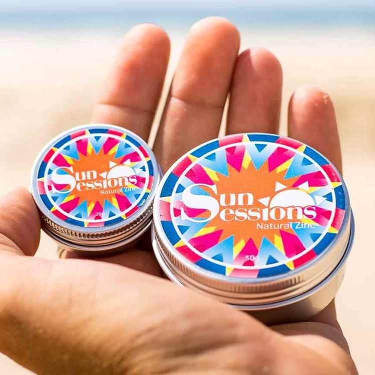 שתי קופסאות קרם הגנה טבעי Sun Sessions Natural Zinc מוחזקות ביד בחוף הים